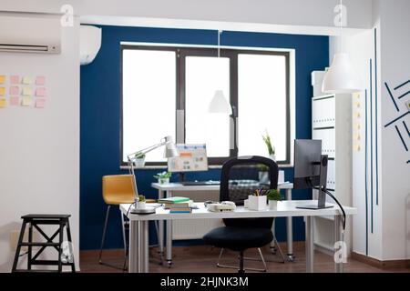 Empty business office room in modern open space workplace. Desk with computer and supplies in commercial workplace with big window. Workspace furniture inside startup with blue and white wall. Stock Photo