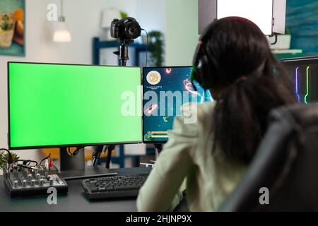 Focused player woman playing online videogames during gaming tournament loooking at mok up green screen chroma key computer with isolated display. Gamer streaming space shooter game using joystick Stock Photo