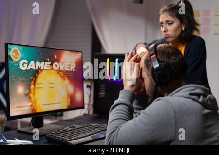Couple of gamers feeling sad about losing video games in front of computer. Woman comforting man about lost game online, having gaming equipment and headphones to play for leisure. Stock Photo