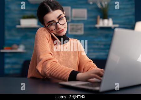 Student with glasses talking on the phone working from home. Programmer focused on working on laptop while using smartphone. Smiling freelancer interacting with technology for remote work. Stock Photo
