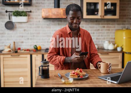 Portrait of smiling black man using smartphone while enjoying breakfast at cozy home kitchen, copy space Stock Photo