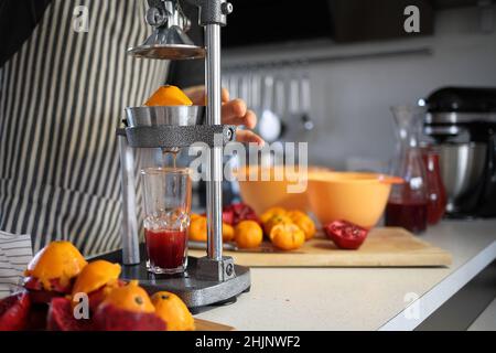 glass of freshly squeezed pomegranate juice, chopped pomegranates and a pomegranate juicer at home. A man in a striped dark apron squeezes juice from Stock Photo