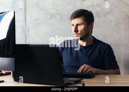 Handsome serious bearded man sitting at the office desk, working on laptop, looking at computer screen against concrete wall in modern office Stock Photo