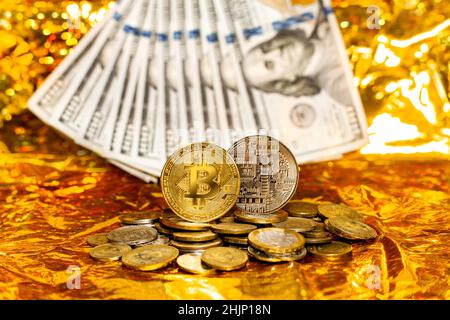 Two bitcoins on a stack of coins against a background of hundred-dollar bills arranged in a fan on a golden background. Stock Photo