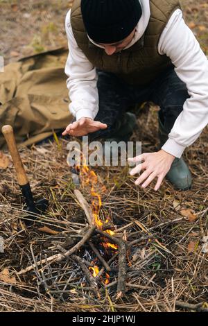 Vertical cropped shot of frozen traveler man warming hands over fire at outdoors on overcast cold day. Stock Photo