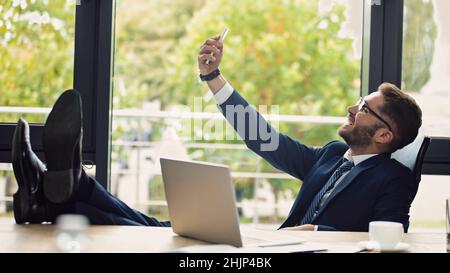 cheerful businessman taking selfie while sitting with legs on desk at workplace Stock Photo