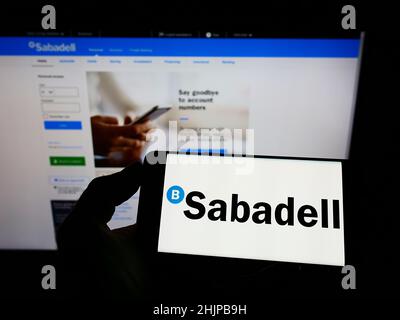 Person holding smartphone with logo of Spanish banking company Banco de Sabadell S.A. on screen in front of website. Focus on phone display. Stock Photo