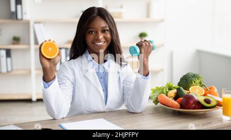 Happy african american woman dietologist holding orange half and dumbbell in her hands, recommending healthy lifestyle Stock Photo