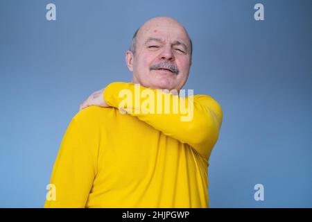 Senior hispanic man suffering from neck, shoulder pain. Studio shot on blue wall. Stock Photo