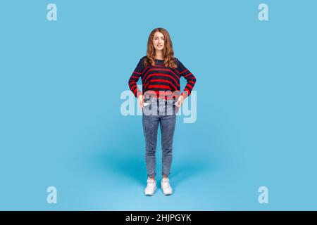 Full length portrait of poor woman, turning out empty pockets, worried about debts, no cash for living, wearing striped casual style sweater. Indoor studio shot isolated on blue background. Stock Photo