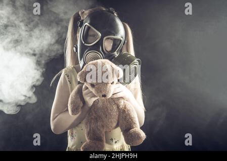Little girl in a gas mask in smoke with a bear in her arms on a black background. Air pollution, respiratory disease. Stock Photo