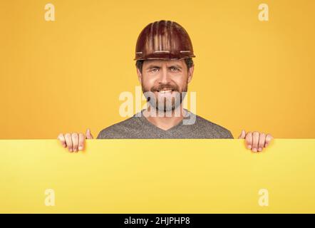 happy mature man builder in helmet behind yellow paper for copy space, advertisement Stock Photo