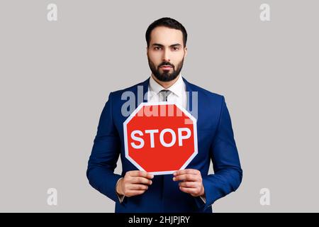 Portrait of serious bearded businessman holding Stop road traffic sign as symbol of prohibition, restrictions, wearing official style suit. Indoor studio shot isolated on gray background. Stock Photo