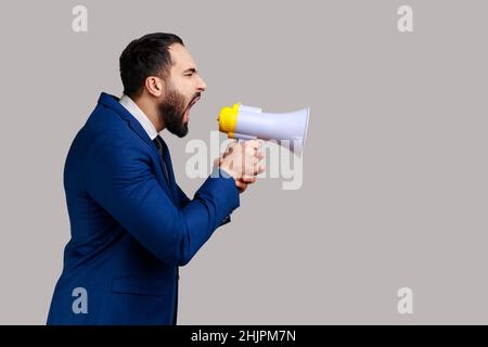 Side view of young adult handsome bearded man screaming in megaphone, announcing important information, wearing official style suit. Indoor studio shot isolated on gray background. Stock Photo