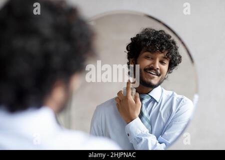 Happy indian businessman in shirt and necktie spraying perfume on his neck while looking and smiling in the mirror Stock Photo