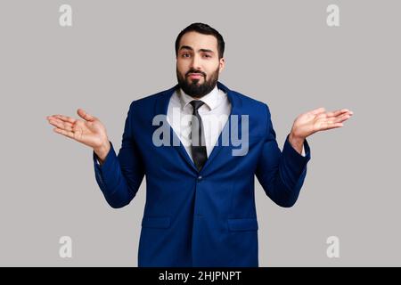 Portrait of confused young adult bearded businessman shrugging shoulders, making no idea gesture, whatever, wearing official style suit. Indoor studio shot isolated on gray background. Stock Photo