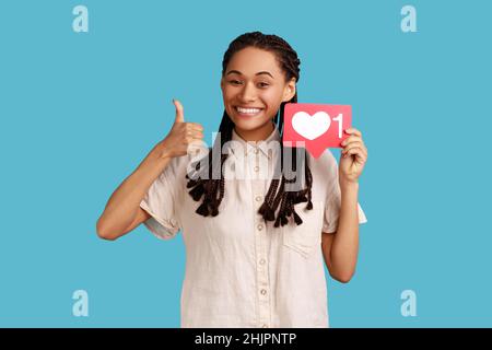 Social media trends. Cheerful woman with black dreadlocks holding network heart Like icon and showing thumb up, recommending to follow excellent blog. Indoor studio shot isolated on blue background. Stock Photo