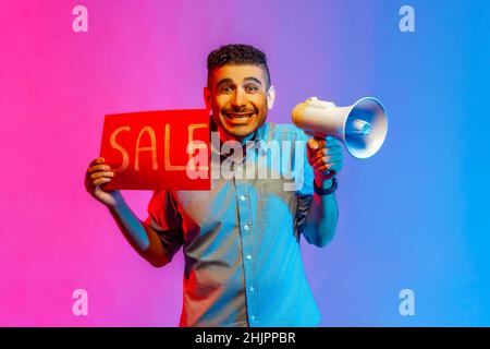 Portrait of smiling happy young adult handsome man in shirt holding megaphone and big sale, black Friday shopping. Indoor studio shot isolated on colorful neon light background. Stock Photo