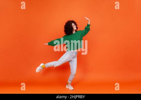 Full length portrait of extremely happy woman with Afro hairstyle wearing green casual style sweater raised arms, posing on one leg, having fun. Indoor studio shot isolated on orange background. Stock Photo