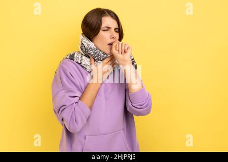 Portrait of unhealthy flu-sick woman standing wrapped in warm scarf shivering from cold, feeling unwell suffering fever, seasonal influenza symptoms. Indoor studio shot isolated on yellow background. Stock Photo