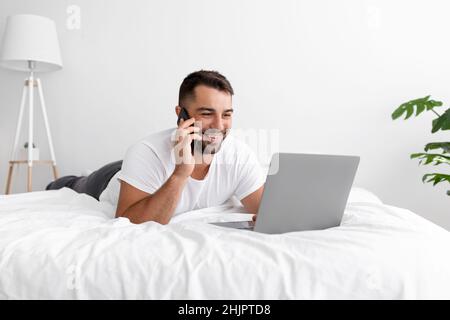 Satisfied caucasian attractive young man in white t-shirt woke up chatting, surfing, working on pc and making phone call Stock Photo