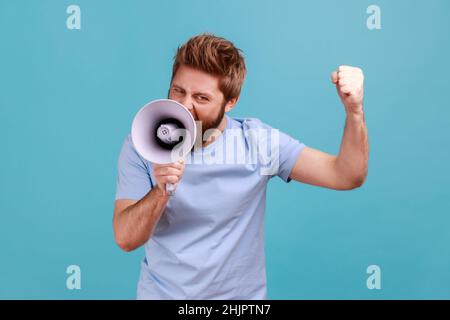 Portrait of aggressive bearded man holding megaphone near mouth, loudly speaking, screaming, making announcement with raised arm, protesting. Indoor studio shot isolated on blue background. Stock Photo