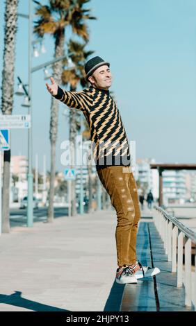 a smiley middle-aged man, wearing hat and modern casual clothes, is about to fall backwards from the bench where is standing in the promenade next to Stock Photo