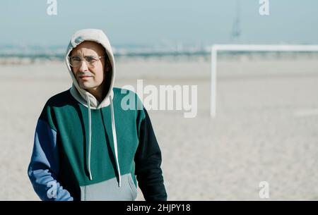 closeup of a mature man, wearing a multicolored hoodie and a pair of modern clear plastic-rimmed eyeglasses, walking by the sand of a urban beach on a Stock Photo
