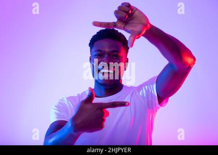 Cheerful young black man making picture frame with fingers, looking at camera and smiling for photo in neon light Stock Photo