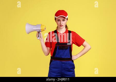 Portrait of strict bossy worker woman holding megaphone in hands, keeps hand on hip, looking at camera with serious expression, wearing overalls. Indoor studio shot isolated on yellow background. Stock Photo