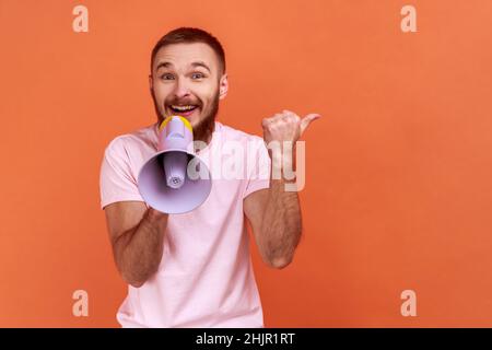 Portrait of bearded man holding megaphone and pointing aside, showing copyspace for advertisement with happy expression, wearing pink T-shirt. Indoor studio shot isolated on orange background. Stock Photo