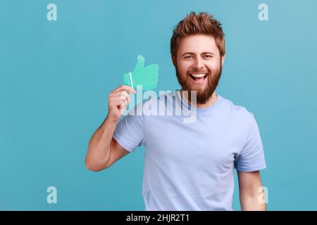 Portrait of extremely happy positive bearded man holding and showing like or thumbs up paper shape sign, looking at camera with toothy smile. Indoor studio shot isolated on blue background. Stock Photo