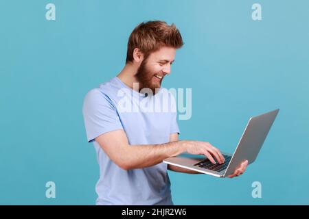 Portrait of smiling bearded man holding laptop in hand and typing, blogger making posts in social networks, chatting with followers. Indoor studio shot isolated on blue background. Stock Photo