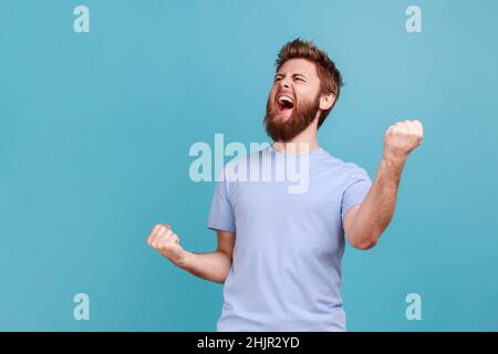 Portrait of overjoyed bearded man standing with excited expression, raising fists, screaming, shouting yeah, celebrating his victory, success. Indoor studio shot isolated on blue background. Stock Photo