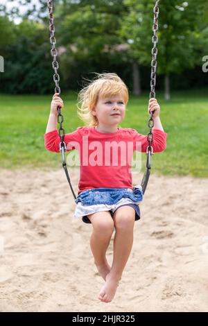 Little girl sitting on a swing in summer. Small beautiful child at playground in park. Stock Photo