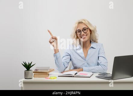 Happy young businesswoman sitting at workplace with pc laptop and pointing finger at free space on light background Stock Photo