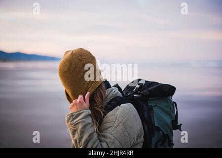 Solo Female Traveler pulling hat on Oregon beach at sunrise Stock Photo