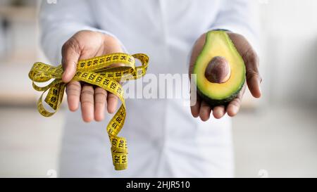 Healthy lifestyle concept. Black female dietitian holding half of avocado fruit and measure tape, closeup, panorama Stock Photo