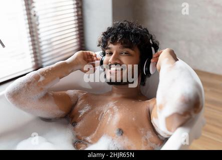 Calm indian man in wireless headphones lying in hot bubbly bath and enjoying his playlist in bathroom at home Stock Photo