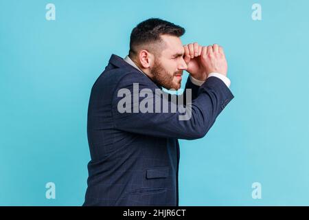 Side view portrait of bearded man wearing official style suit making glasses shape, looking through binoculars gesture with surprised expression. Indoor studio shot isolated on blue background. Stock Photo