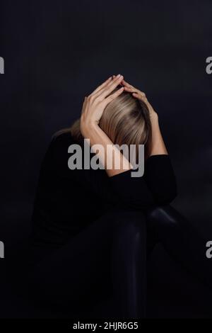 Caucasian woman in depression wearing black dress being on floor hiding face with arms in despair with black background. Mental and physical abuse Stock Photo
