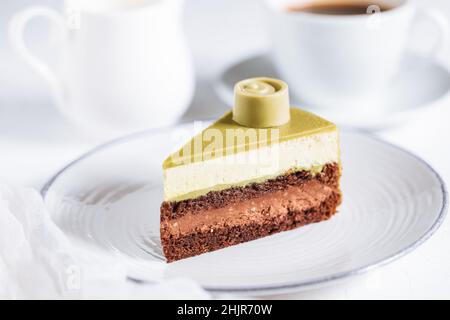 Pistachio chocolate cake with four layers and green pistachio glazing on top served on a white plate on white background with coffee in blur Stock Photo
