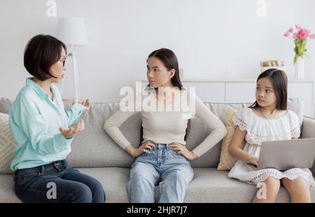 Little Asian girl with laptop computer and young woman having conflict with mature lady, sitting on couch at home Stock Photo