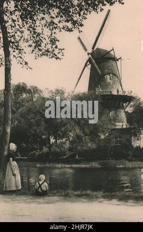 This giant windmill grinds the corn brought from the farms round Middelburg along the gently flowing canal at its foot. Netherlands. Holland (1923) Stock Photo