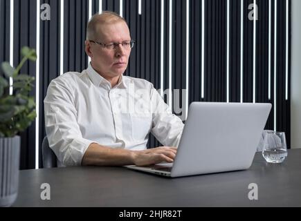 Concentrated and serious mature businessman in glasses sitting at table and typing on laptop keyboard. Employee working in office. High quality photo Stock Photo
