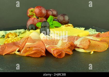 Assorted cheeses on black slate board. Brie cheese, cheddar slices, walnuts, grapes, olives and Schwarzwald ham. Still life food. Copy space Stock Photo