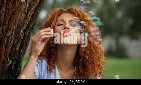 curly redhead woman frowning while blowing soap bubbles in park Stock Photo