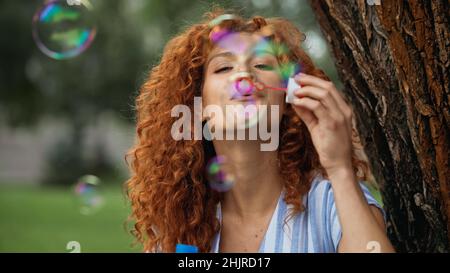 cheerful redhead woman blowing soap bubbles in park Stock Photo