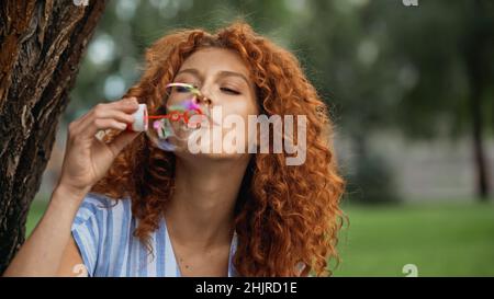 curly redhead woman blowing soap bubble in park Stock Photo