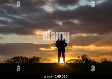 Rear view silhouette of isolated woman in winter coat & wellies on countryside walk looking at sunset sky, watching sun setting with sunburst effect. Stock Photo